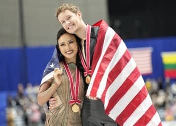 Gold medalists Madison Chock and Evan Bates of the United States side hug as they skate during the victory ceremony at NHK Trophy. They both wear gold medals, the U.S. flag draped around them as they smile.